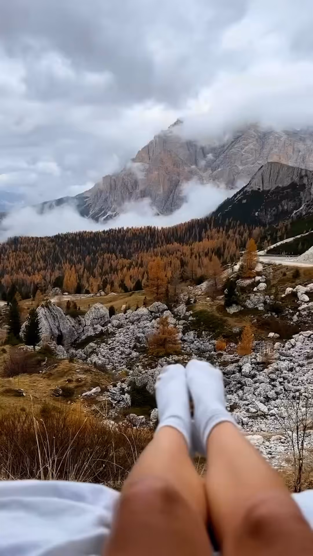 Person lying down with legs extended, overlooking a rocky mountain landscape with autumn trees and low clouds.
