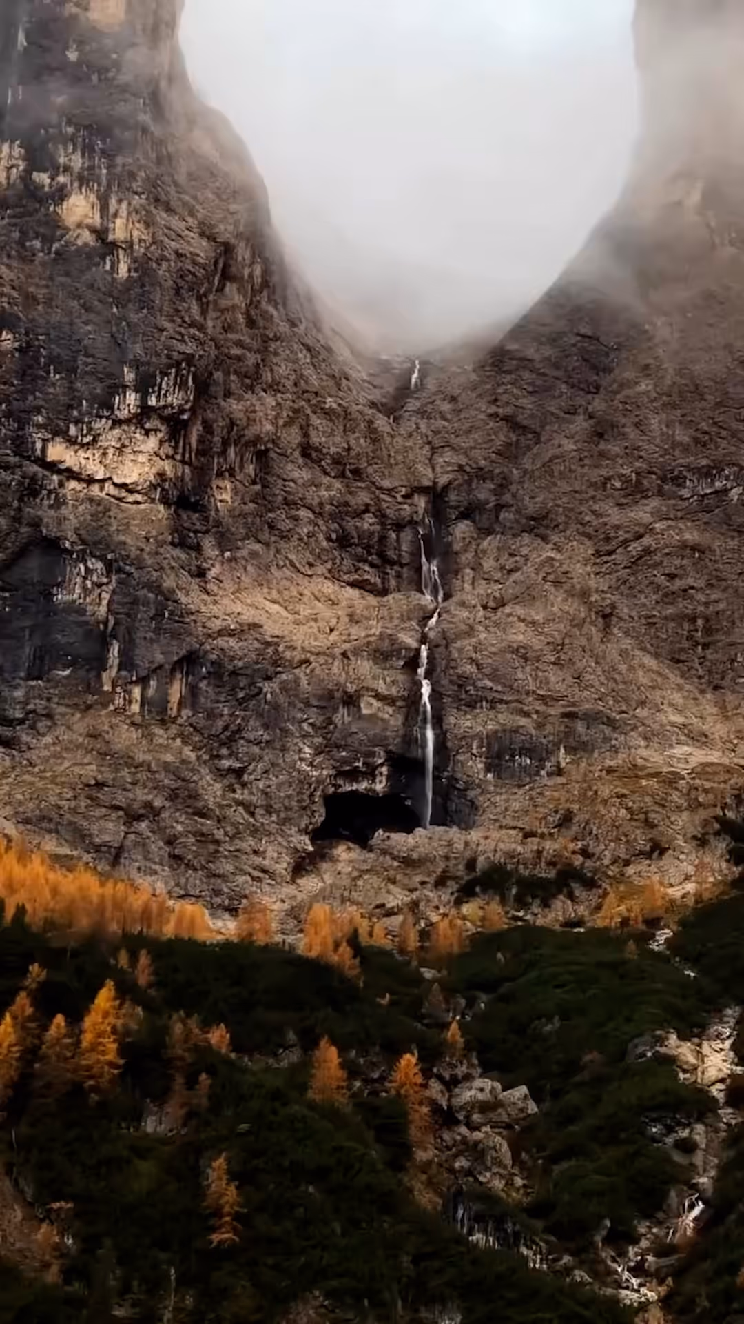 Tall rocky mountain with a narrow waterfall cascading down from a misty peak and orange and green trees at the base.