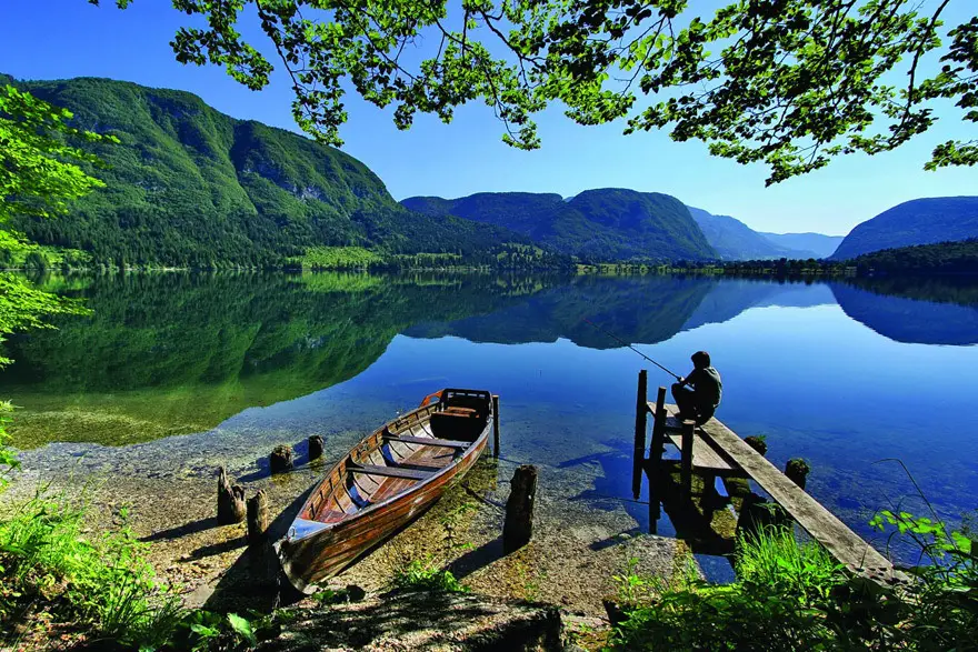 Person fishing from a wooden dock beside a wooden boat on a clear lake with green mountains reflected in the water under a blue sky.