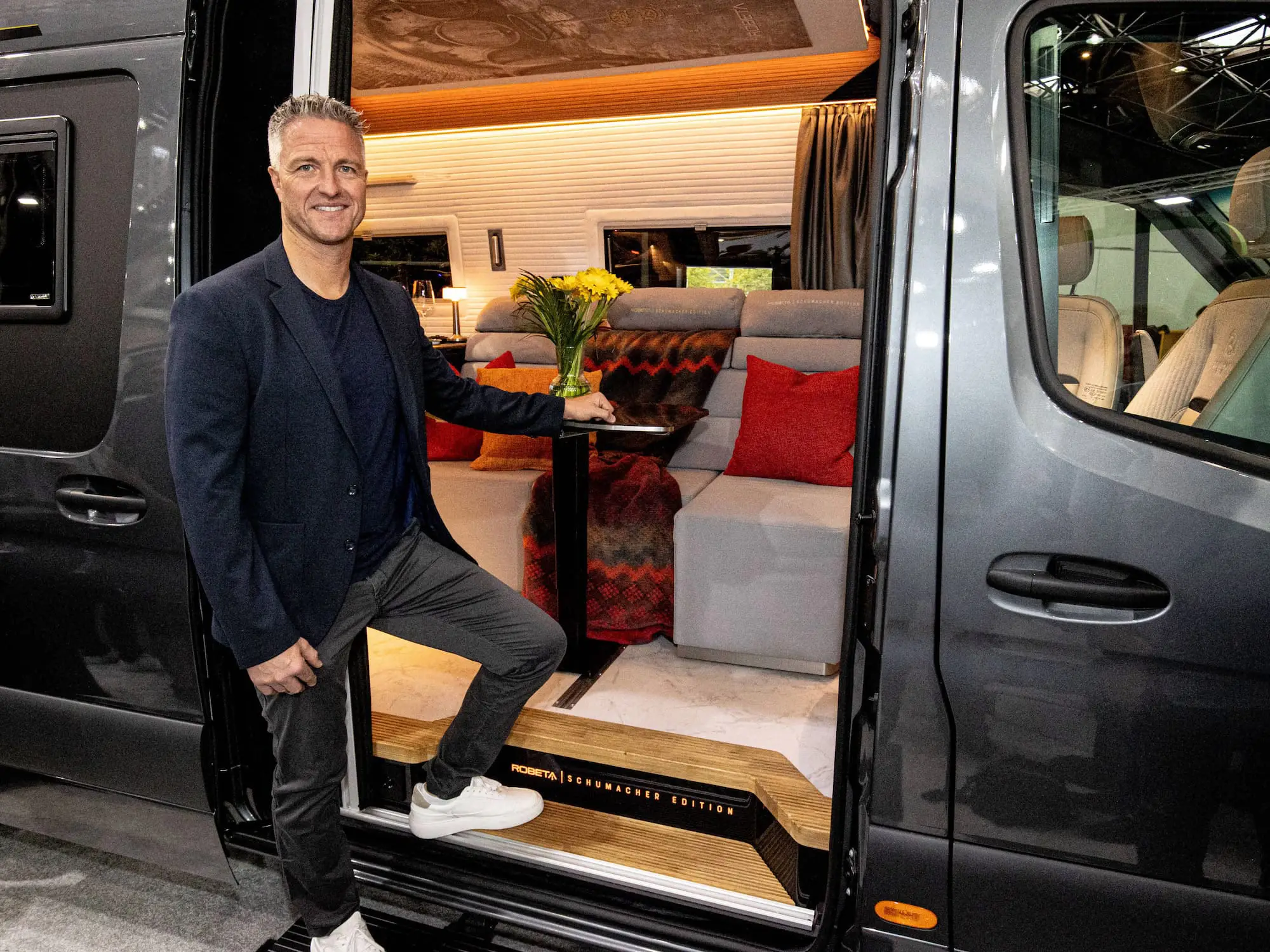Man in a dark blazer and white sneakers standing at the open door of a gray van, showing the interior with gray seating, red cushions, a small table with yellow flowers, and a sign that reads 'Robeta Schumacher Edition'.