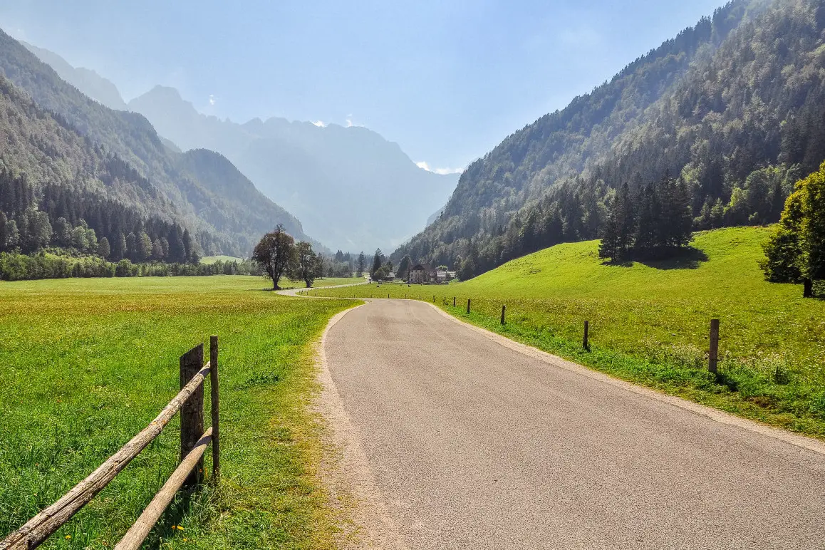 Paved road winding through a green valley surrounded by forested mountains under a clear blue sky.