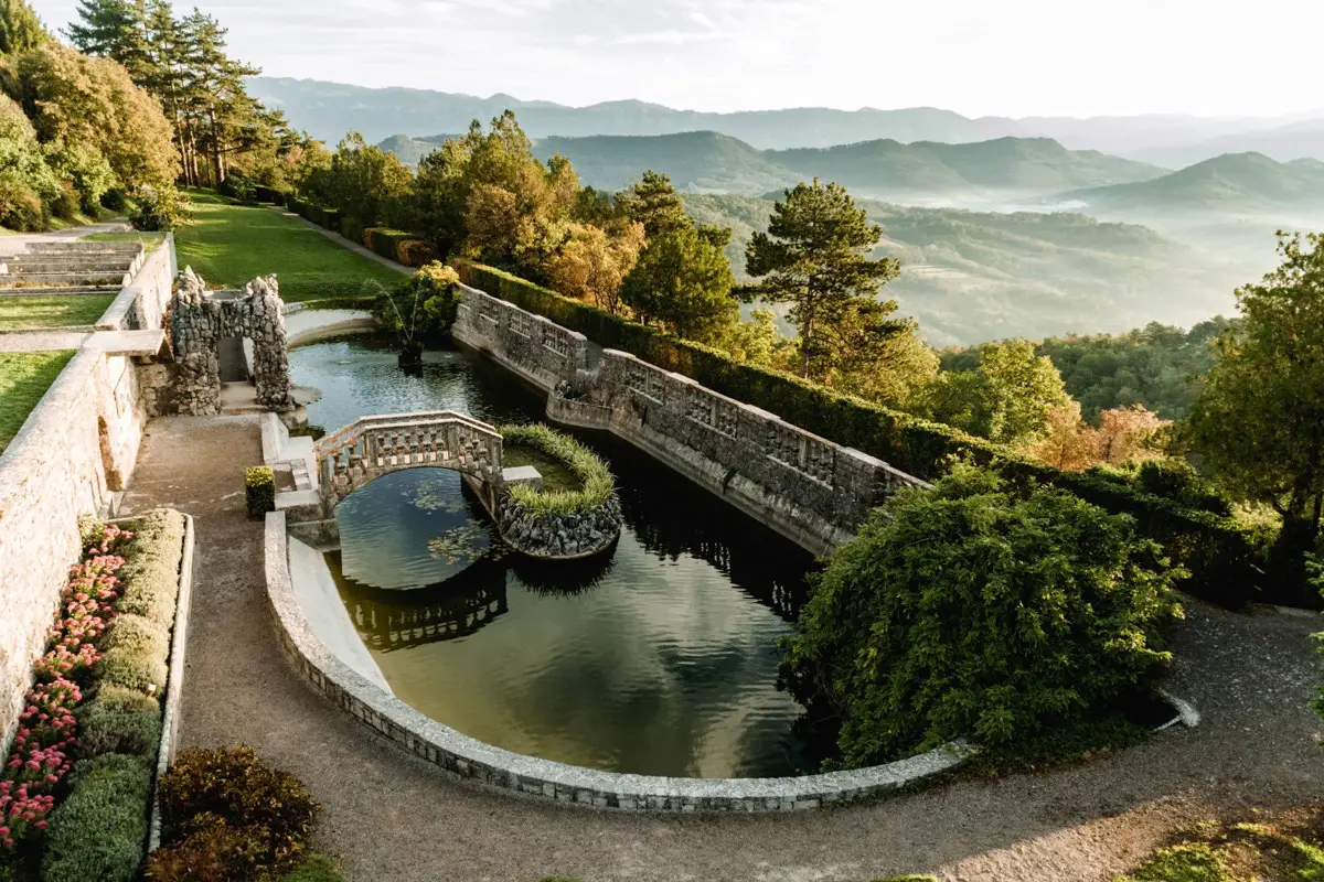 Stone bridge over a decorative pond in a terraced garden with trees and mountains in the background.