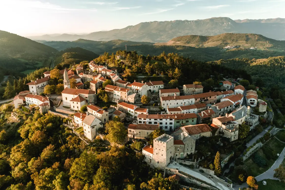 Aerial view of a historic village with terracotta rooftops surrounded by dense trees and hills under a bright sky.