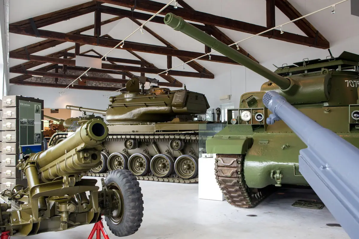 Interior of a military museum featuring green tanks and artillery pieces under wooden ceiling beams.