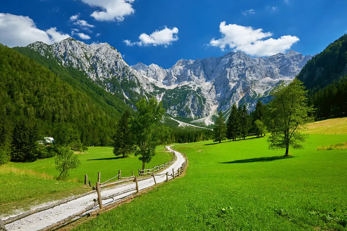 A winding dirt path bordered by wooden fences runs through green meadows with scattered trees, leading to steep rocky mountains under a blue sky with scattered white clouds.