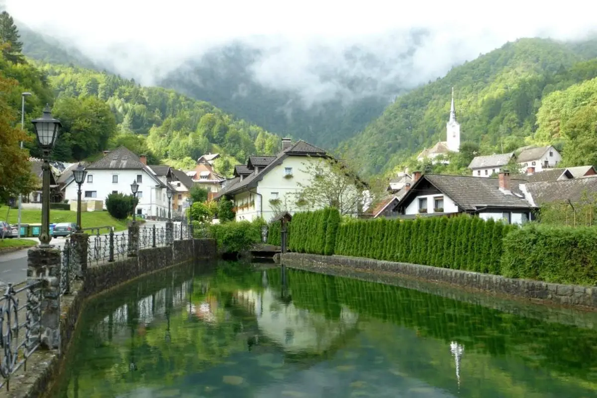 A peaceful village with houses and a church reflected in a calm river, surrounded by green forested mountains under misty sky.