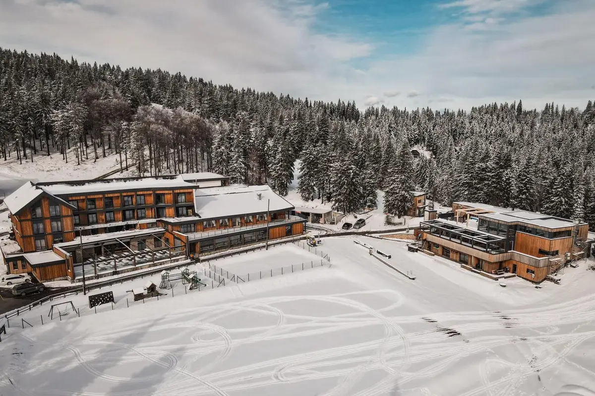 A wooden lodge surrounded by snow-covered ground and dense snow-laden pine trees under a partly cloudy sky.