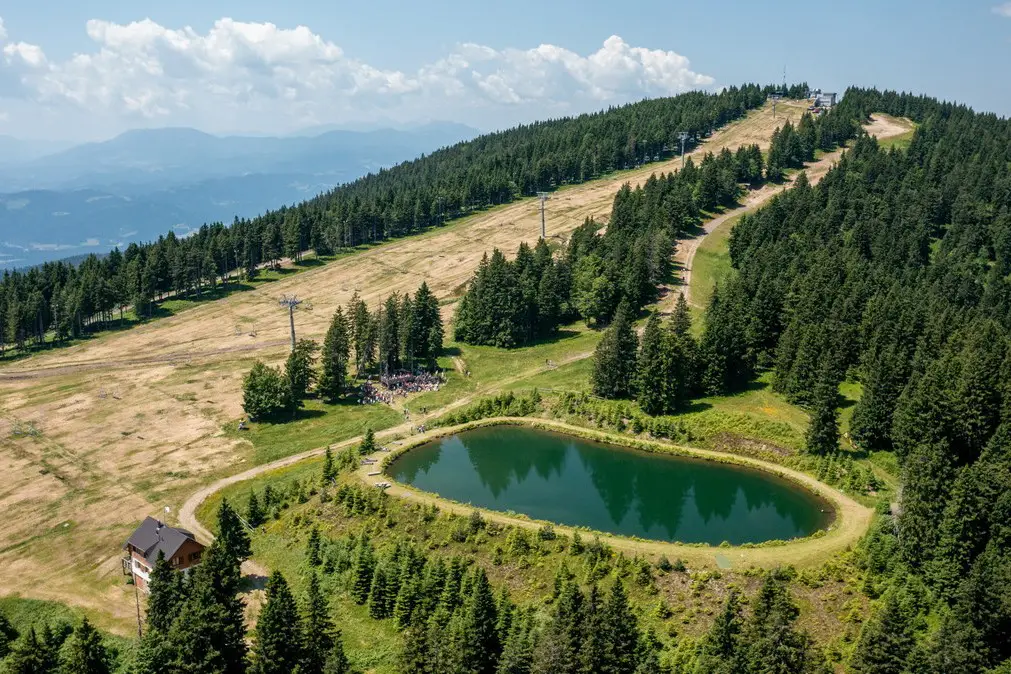Aerial view of a mountain landscape with a small pond, grassy slopes, scattered trees, a cabin, and ski lift infrastructure.