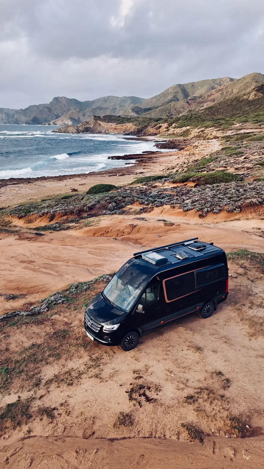 Black camper van parked on sandy terrain near a coastal rocky beach with waves and distant mountains under a cloudy sky.