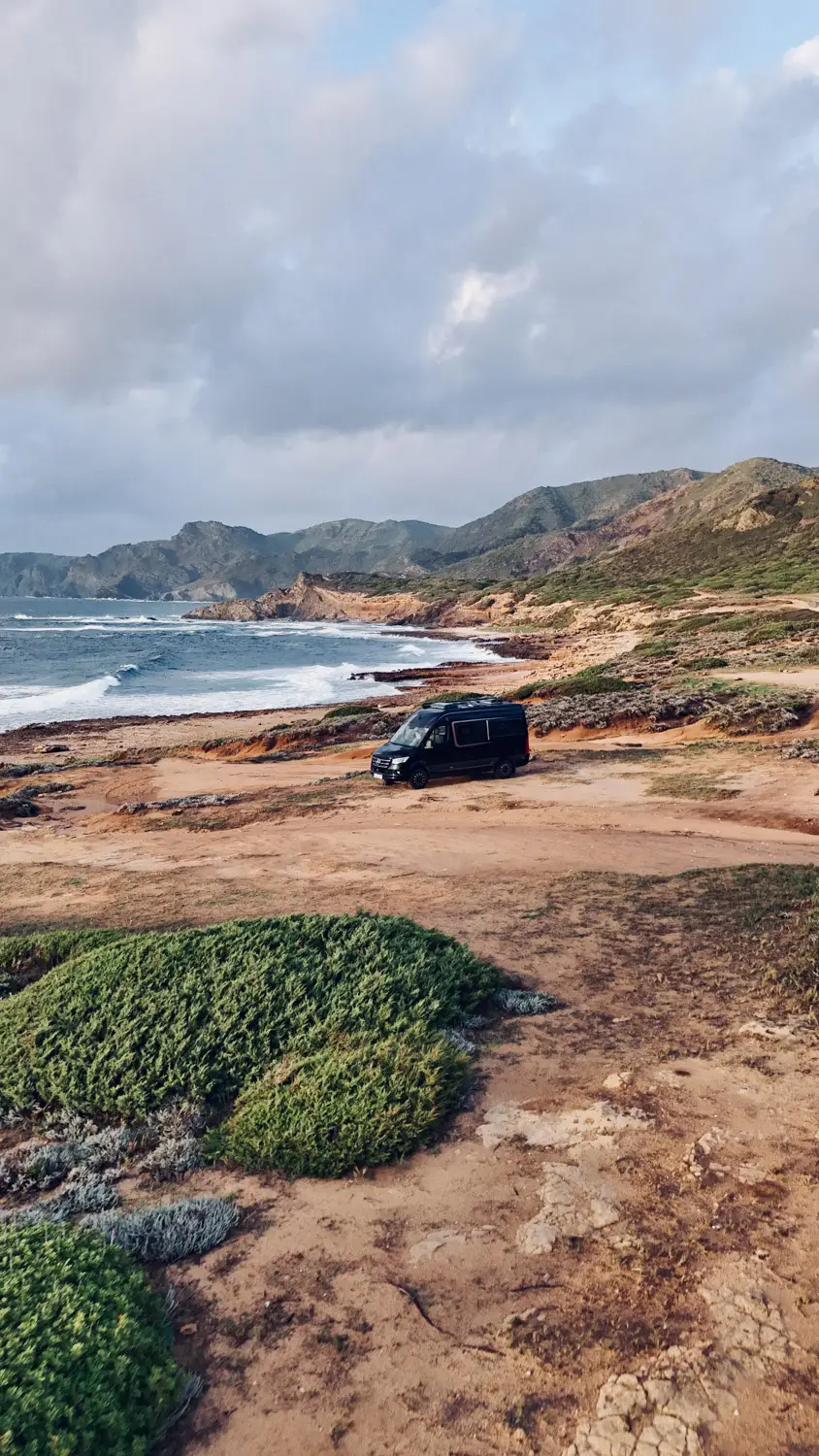 Black camper van parked on a rocky coastal landscape with green shrubs and mountains in the background under a cloudy sky.