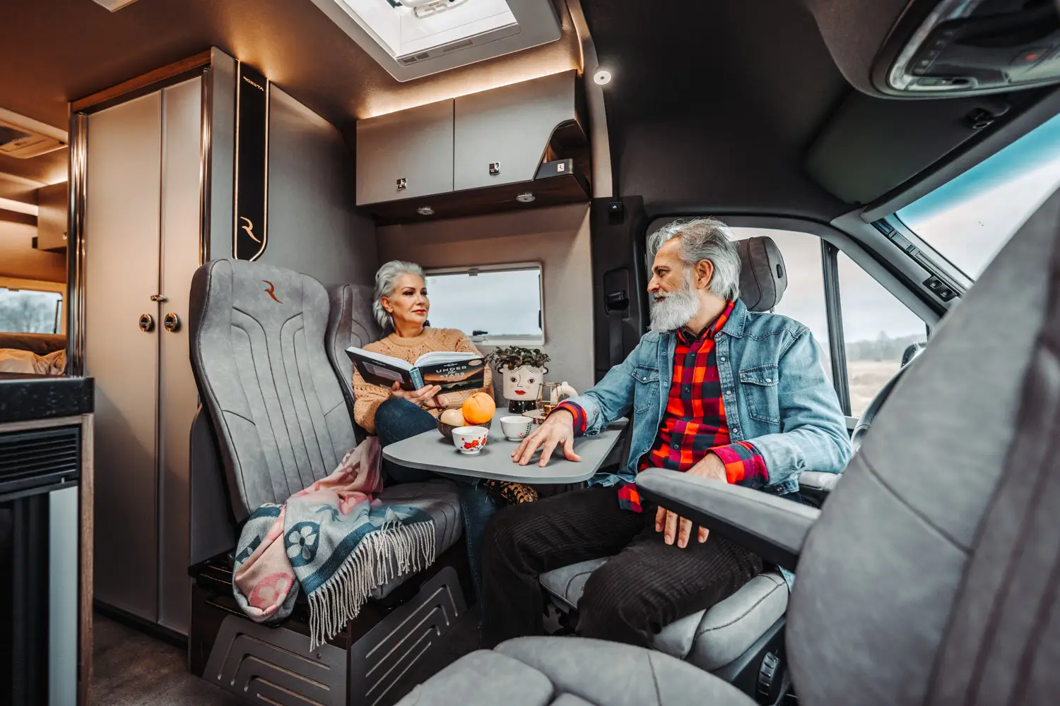 Older couple sitting inside a camper van, woman reading a book and man looking at her, with a small table holding cups and fruit between them.