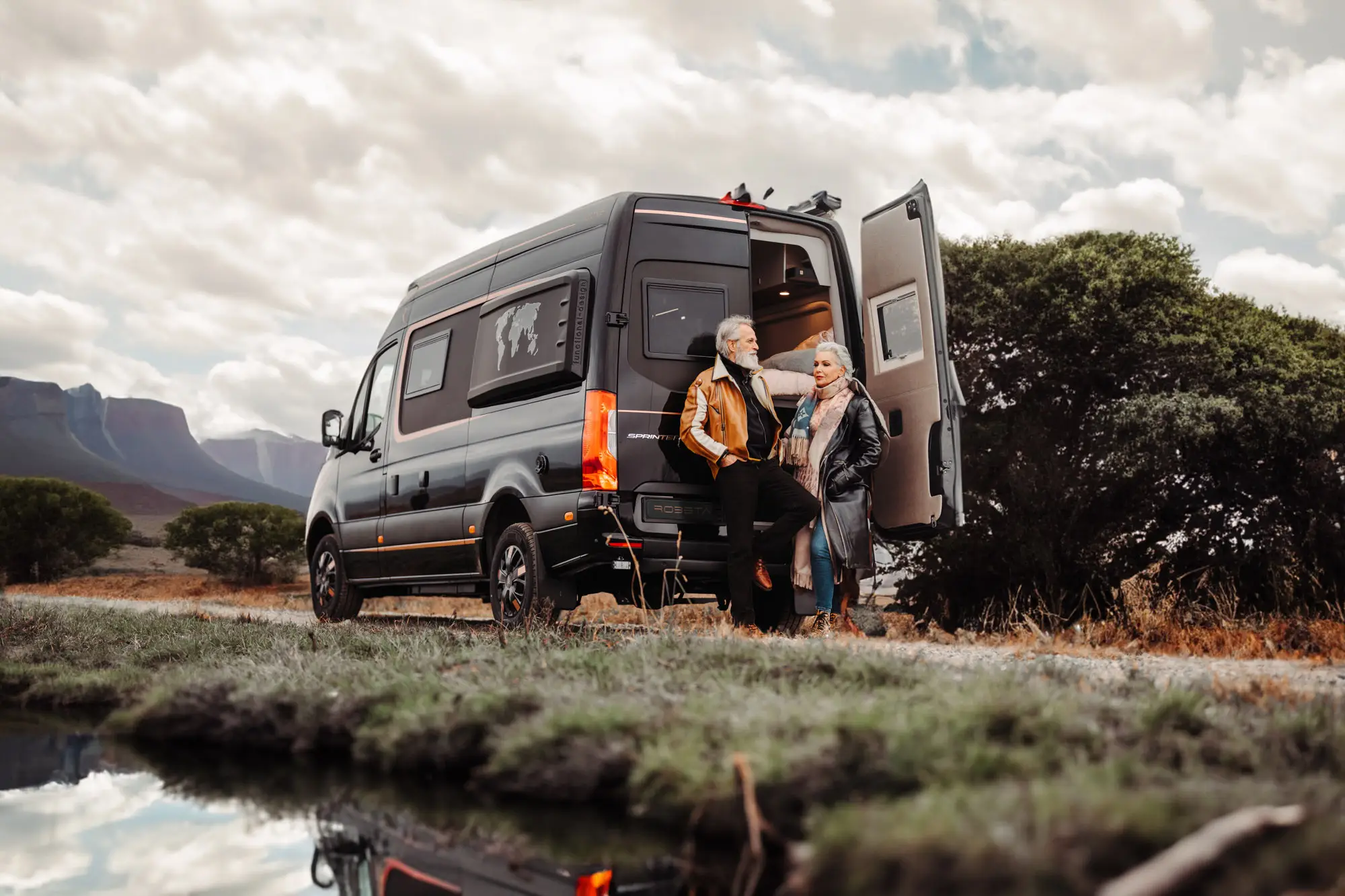 Older couple standing by the open back door of a black camper van parked in a scenic outdoor area with mountains and trees.