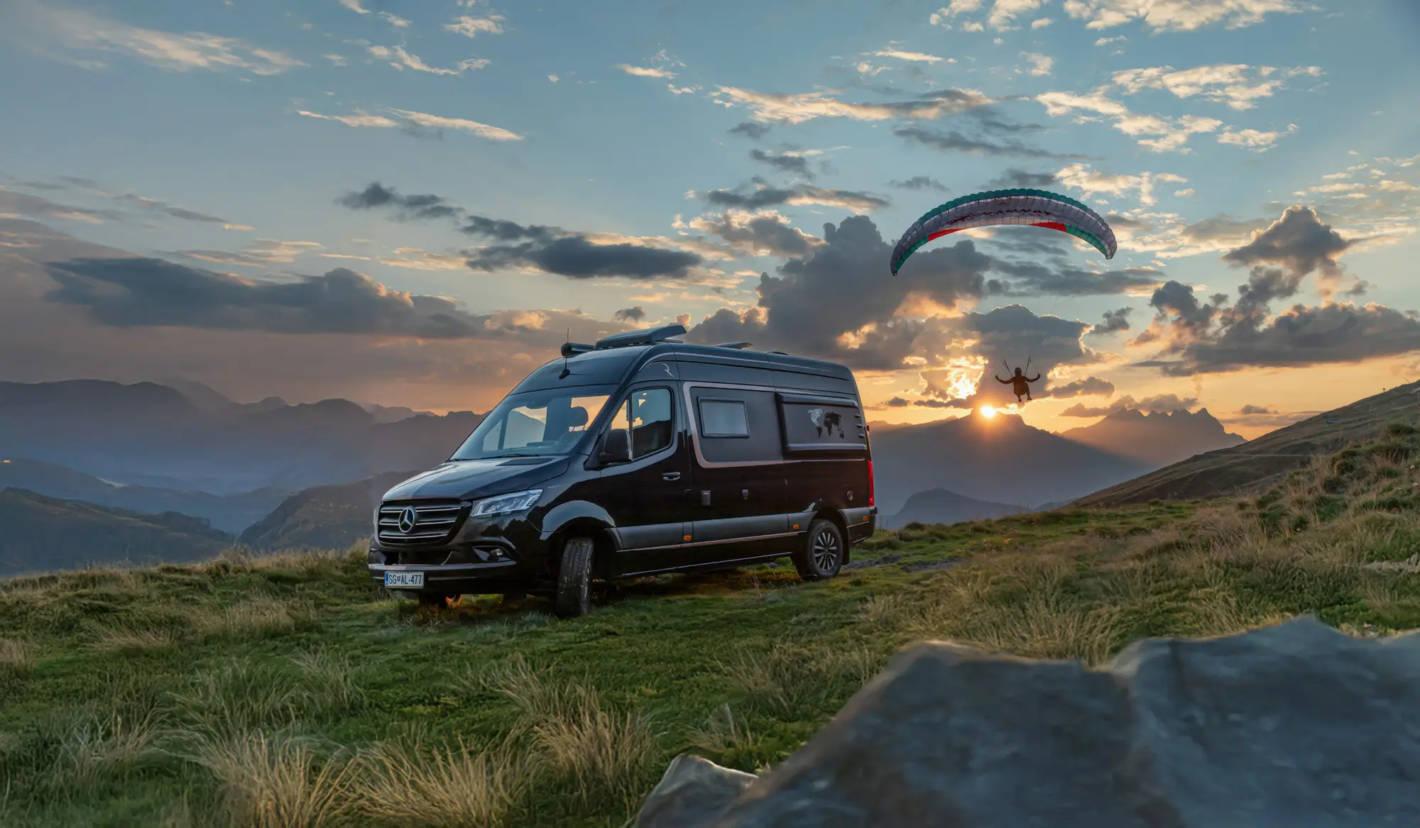 Black camper van parked on grassy hillside at sunset with a paraglider flying nearby against a backdrop of mountains and clouds.