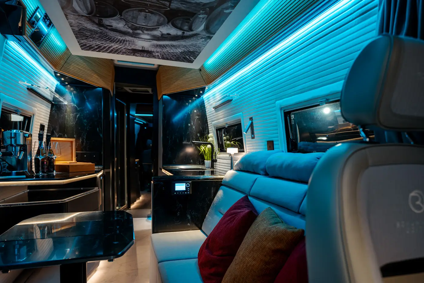 Luxurious modern interior of a camper van with LED lighting, black marble surfaces, beige seating with colorful pillows, and a small kitchenette.