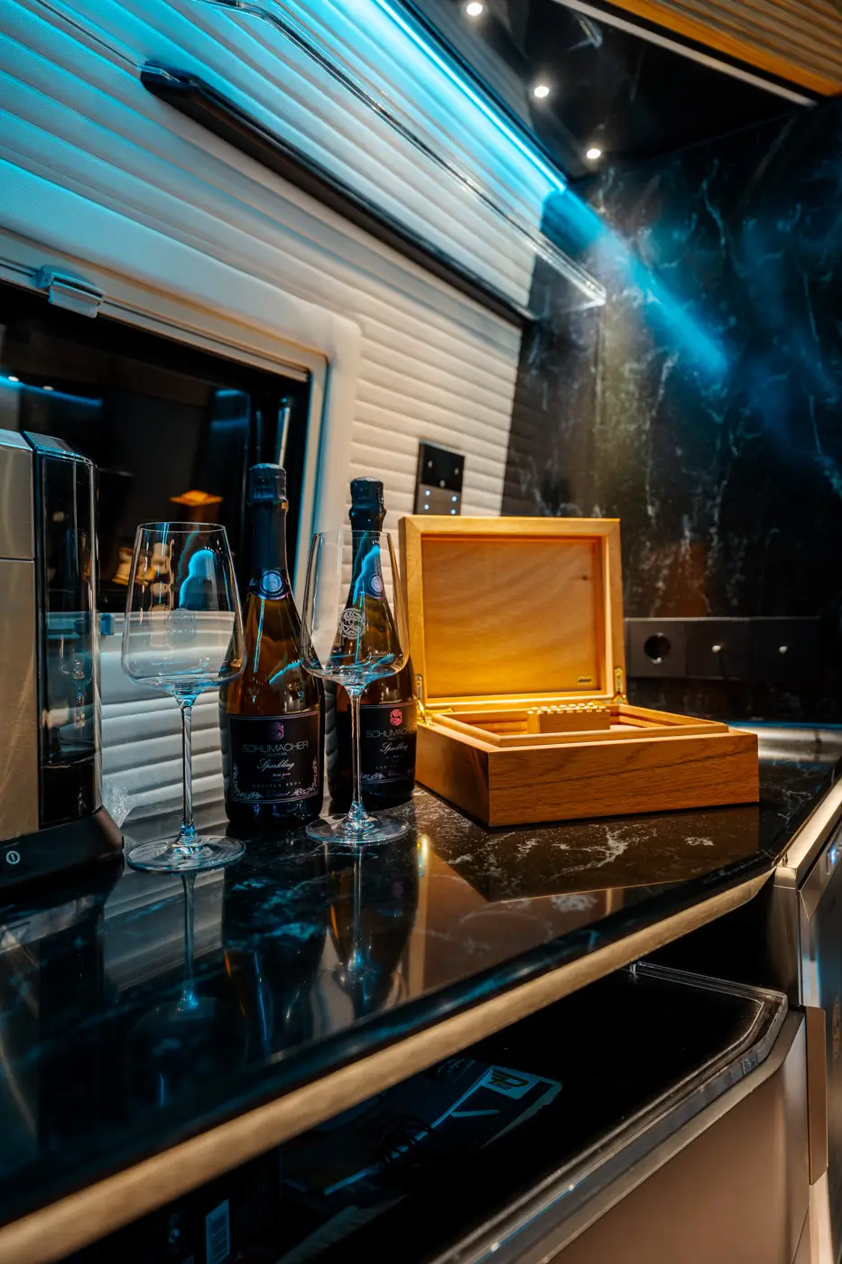 Two bottles of Schumacher sparkling wine, two empty wine glasses, and an open wooden box on a black marble countertop with blue lighting.