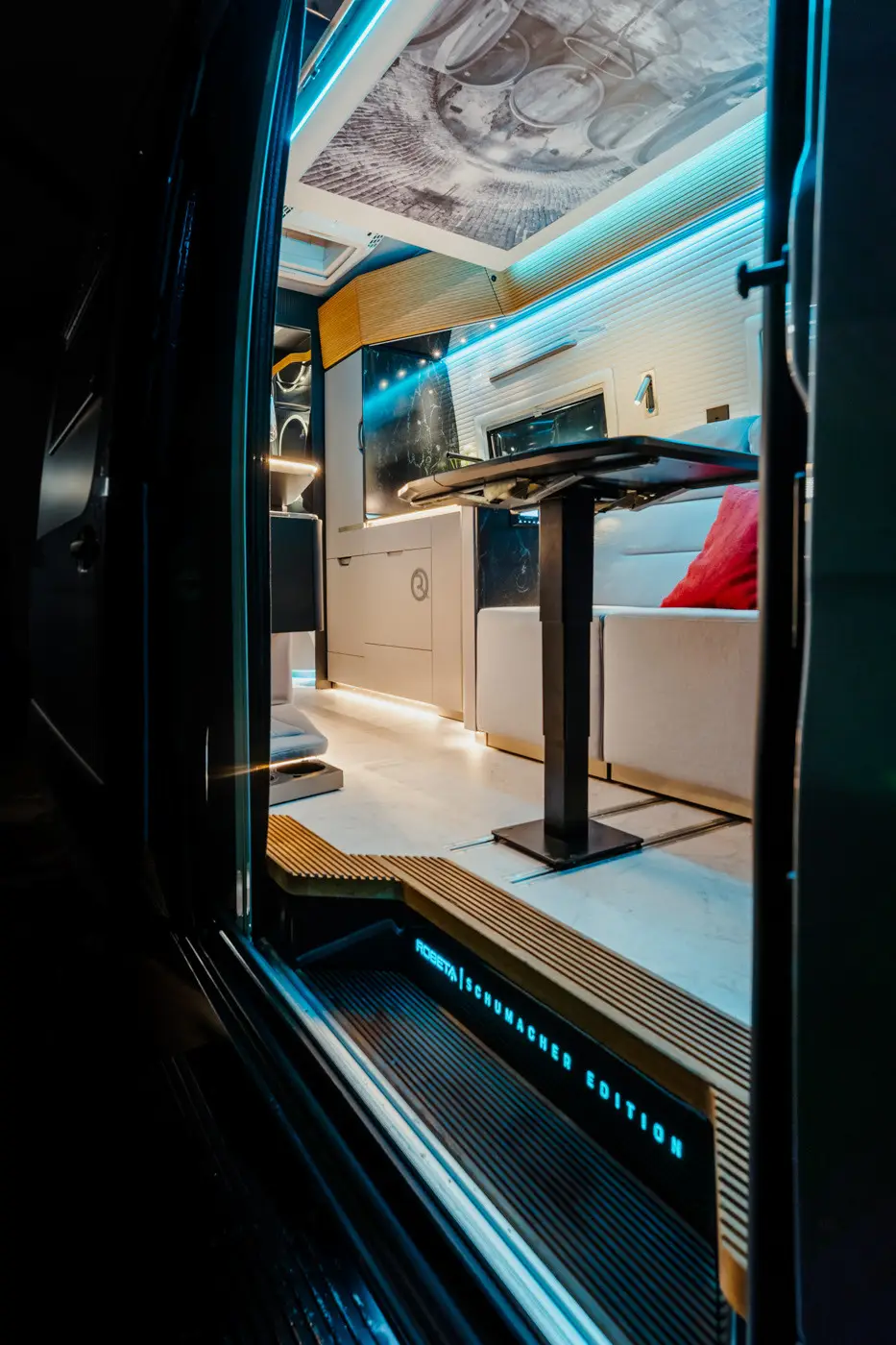 Interior view of a modern camper van featuring a dining table, white cushioned bench with a red pillow, and ambient blue lighting.