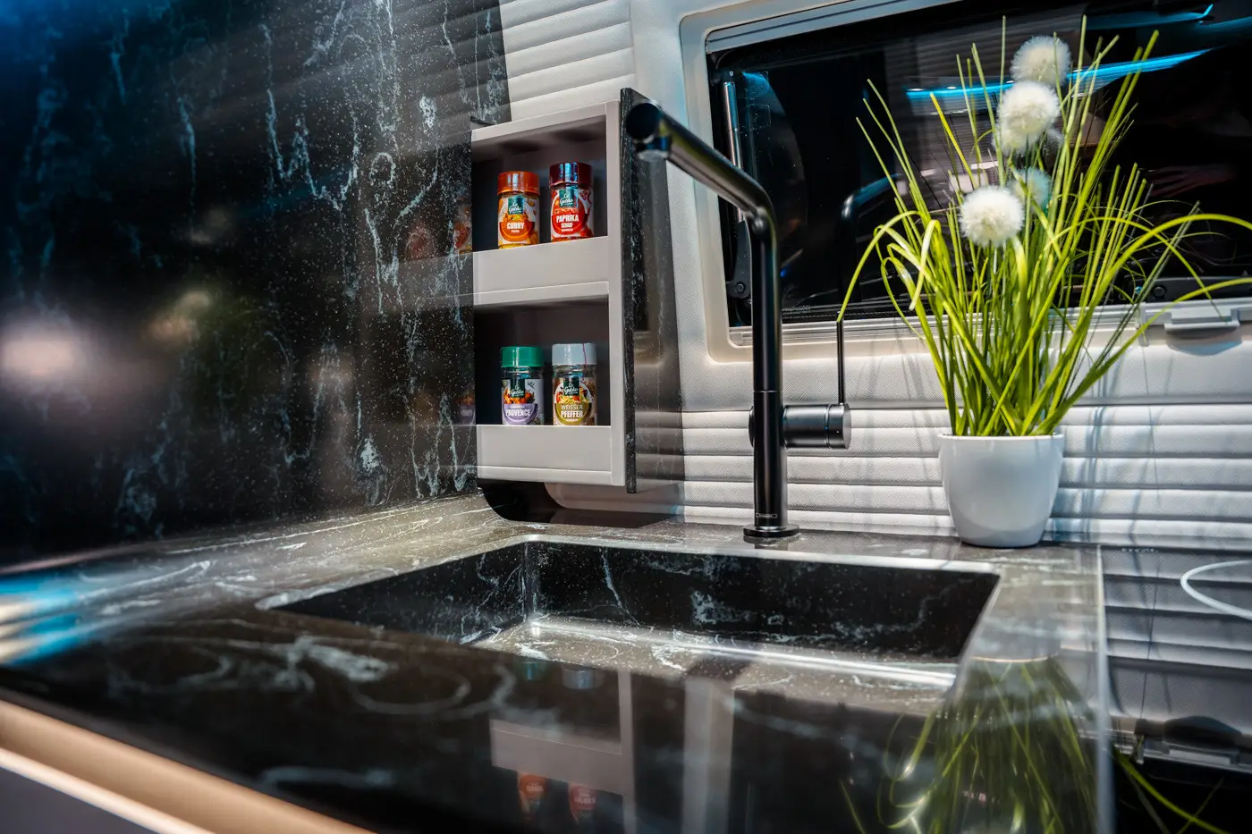 Modern kitchen sink with black marble countertop, black faucet, spice jars on a white shelf, and a white pot with decorative green grass and white pom-poms.
