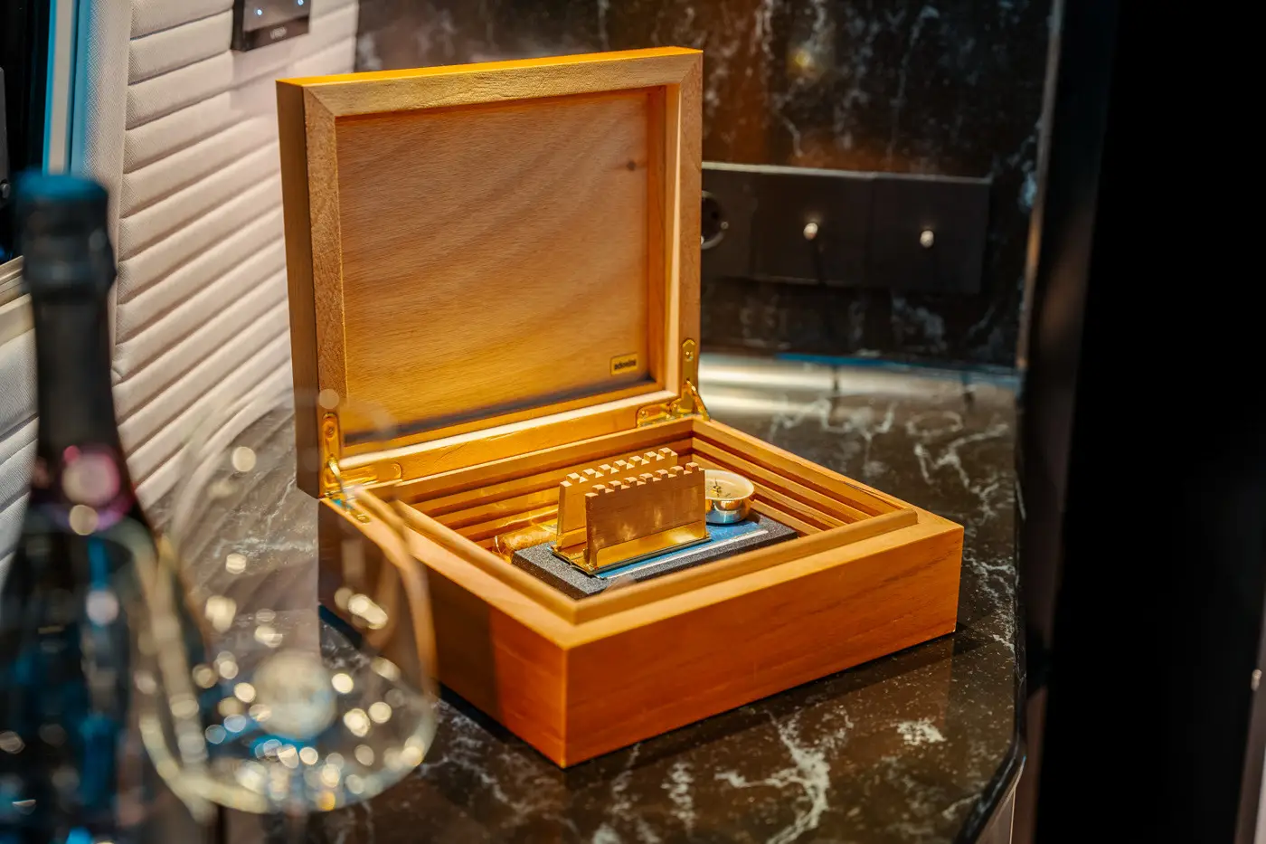 Open wooden humidor box on a marble countertop with a cigar and hygrometer inside, with a blurred wine bottle and glass in the foreground.