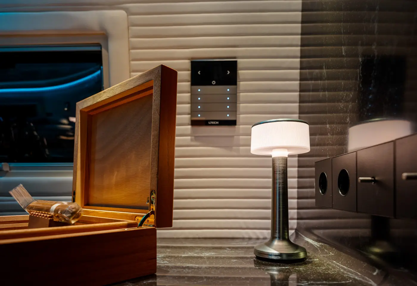 Modern interior scene with an open wooden cigar box containing a cigar, a lit metal and white lampshade table lamp on a marble surface, and a control panel on a ribbed wall.