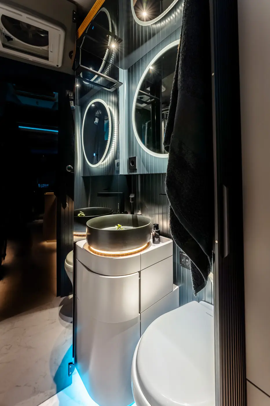 Modern bathroom interior with a round illuminated mirror, a black vessel sink on a white vanity, and a white toilet.