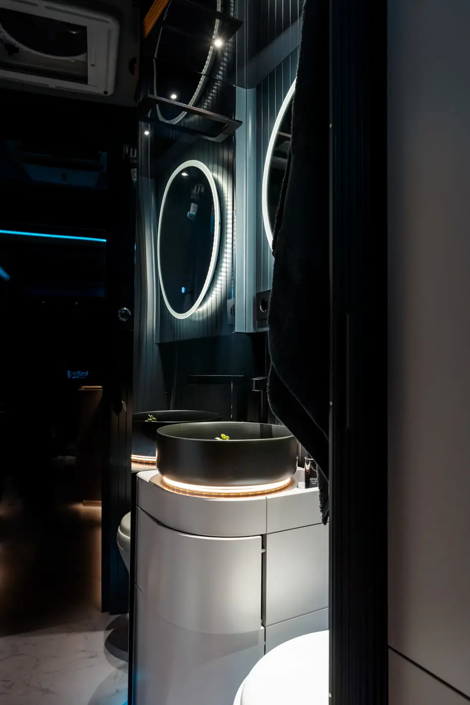 Modern bathroom with a round black vessel sink illuminated by LED strip lighting under it, a circular backlit mirror, and dark vertical paneling.