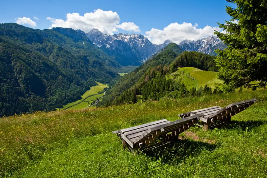 Two wooden lounge chairs on a grassy hillside overlooking green mountains and a partly cloudy blue sky.