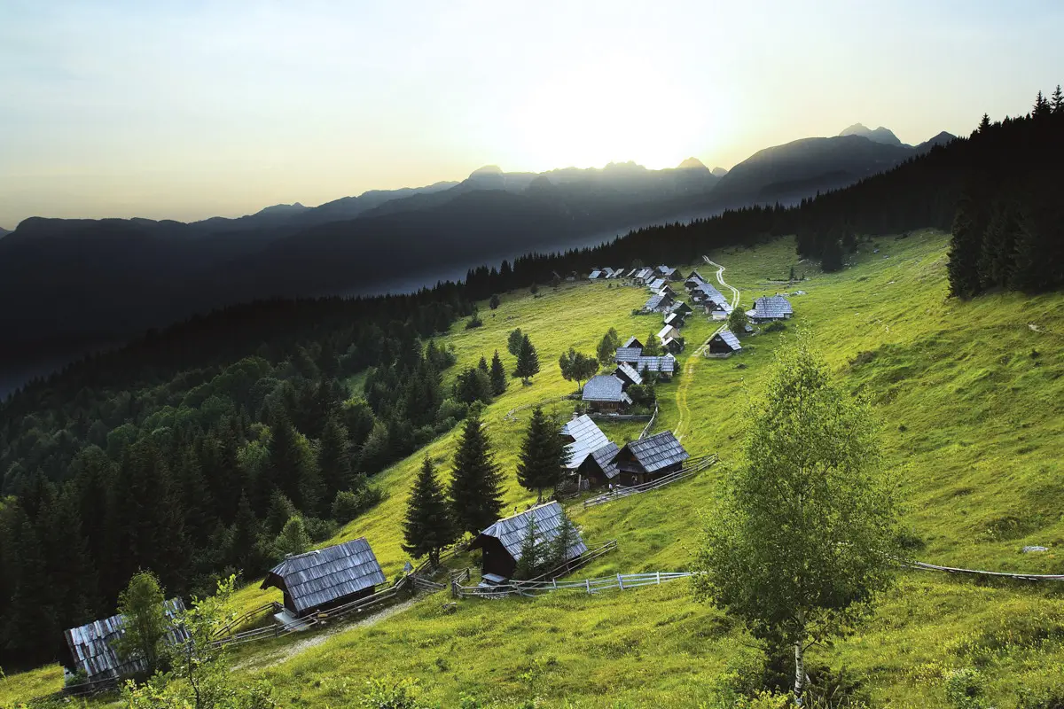Small mountain village with wooden cabins scattered across a green hillside under a setting sun.