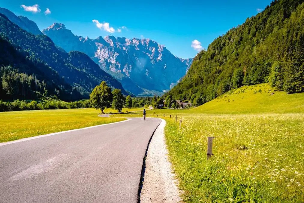 A winding road through green fields with a cyclist riding toward distant rocky mountains under a clear blue sky.