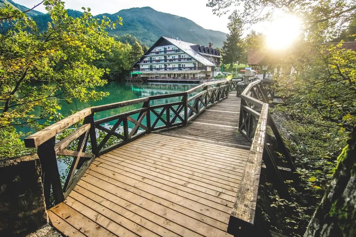 Wooden bridge over clear green lake leading to a large house with mountains in the background and sunlight filtering through trees.