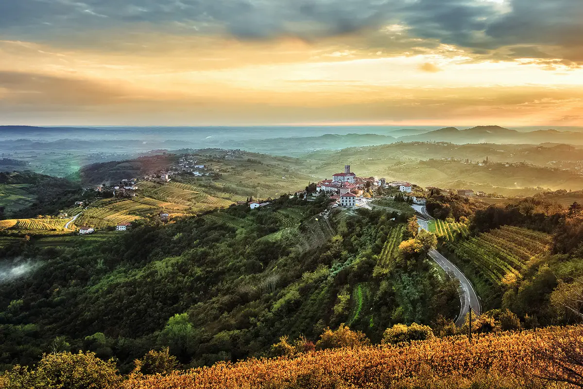 A winding road leading to a small hilltop village surrounded by lush green fields and forests under a cloudy sunset sky.
