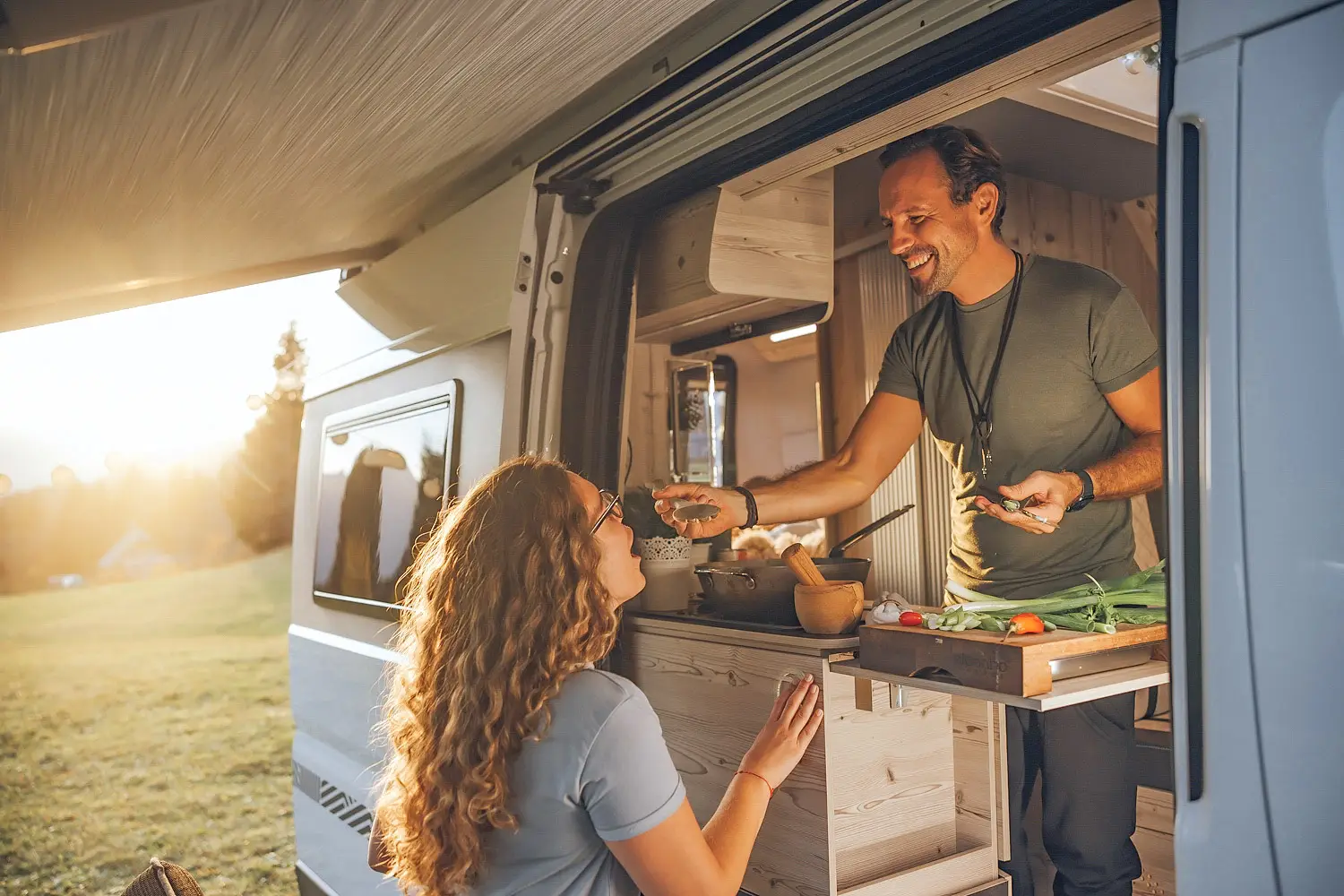 Man feeding woman by hand from the kitchen window of a camper van at sunset.