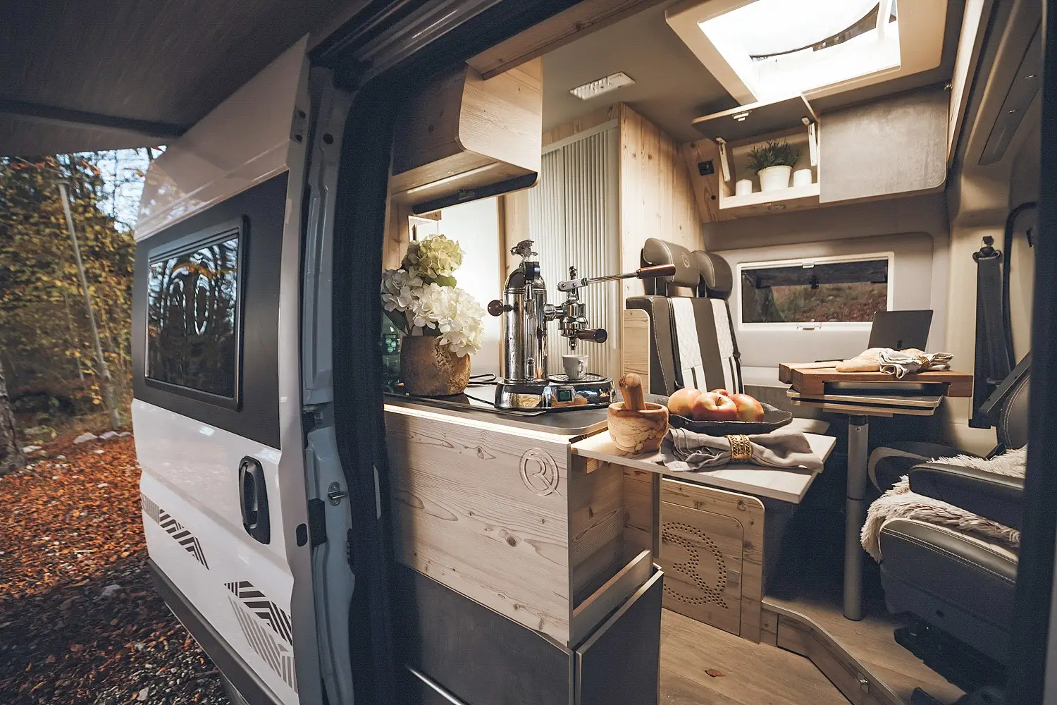 Interior of a camper van with wooden cabinetry, a coffee machine, a tray of apples, a laptop on a table, and cushioned seats.