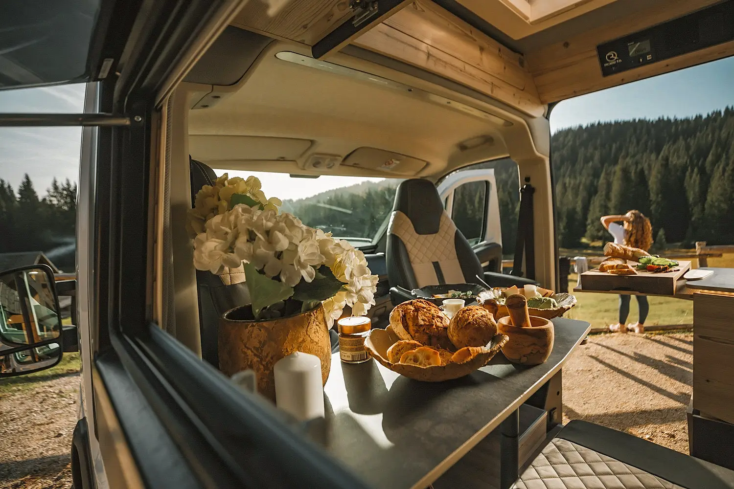 Interior of a camper van with a table set with bread, a plant, and food, with a woman standing outside in a sunny forested area.