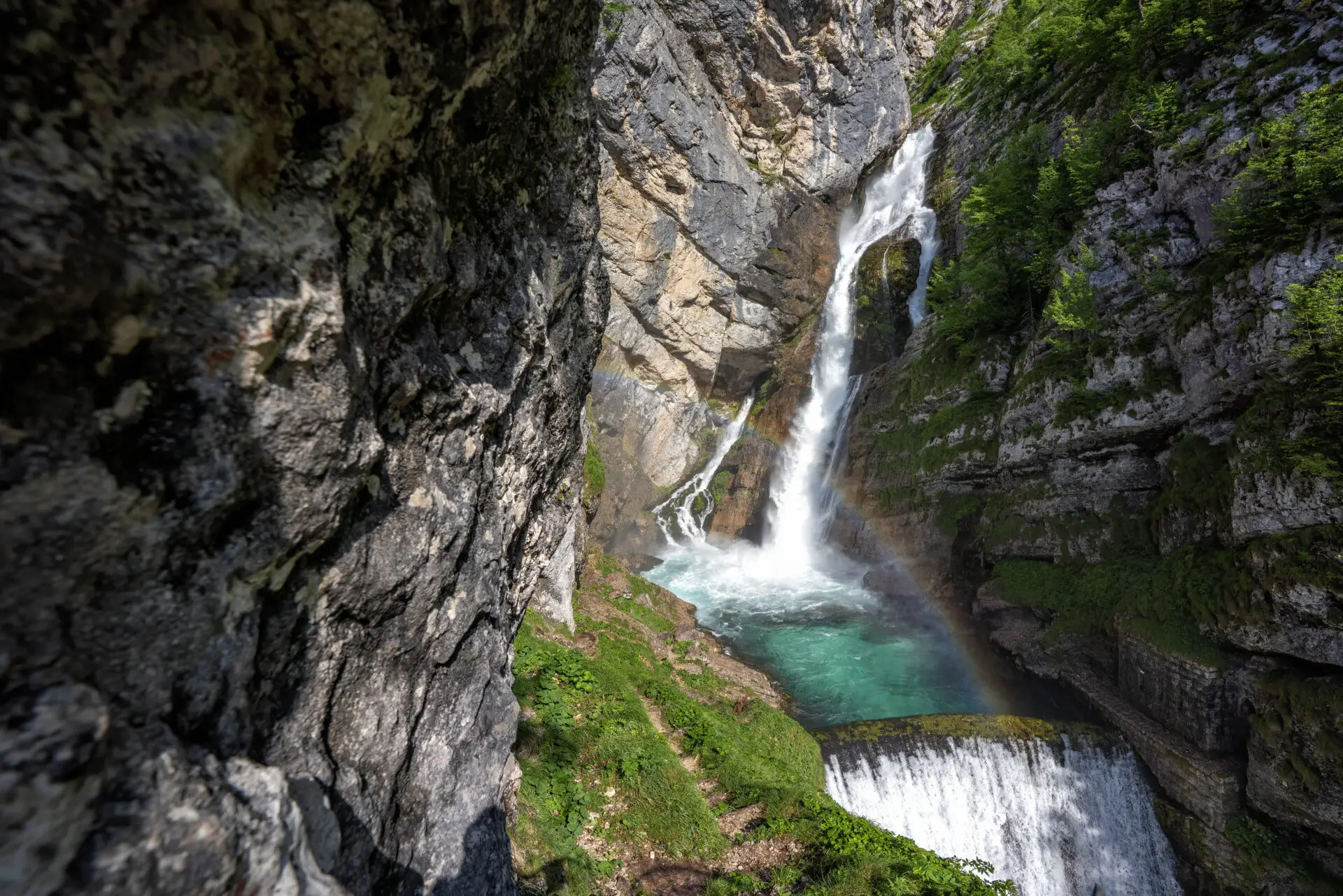 Tall waterfall cascading into a turquoise pool surrounded by rocky cliffs and green vegetation with a rainbow arcing through the mist.